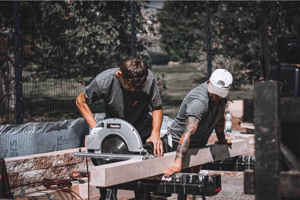 Zwei Dachdecker arbeiten im Team und schneiden auf der Baustelle einen Holzbalken mit der Kreissäge präzise zu